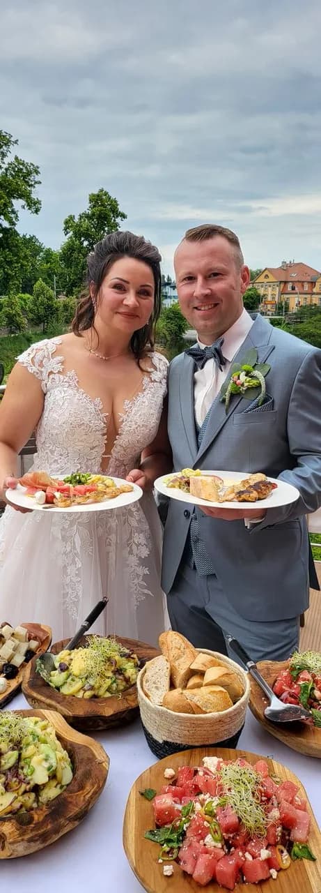 Ein Brautpaar hält Teller mit Vorspeisen, davor ein Buffet mit Salaten und Brot